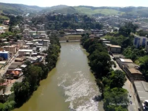 Imagem aérea de um trecho da cidade de Ponte Nova. No centro, a extensão do Rio Piranga, com o pontilhão de ferro ao fundo e a beira rio ao lado.